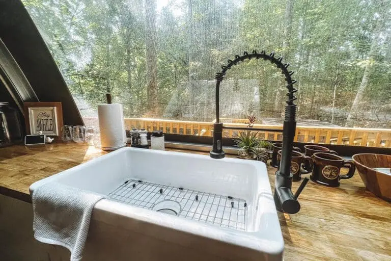 Rustic farmhouse kitchen sink with black gooseneck faucet overlooking a lush forest through a large window.