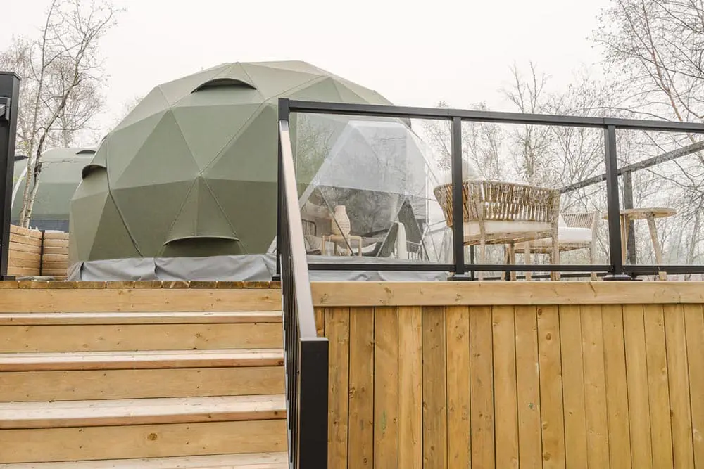 Geodesic dome outdoor glamping pod on wooden deck with seating area and glass railing, natural wooded surroundings.