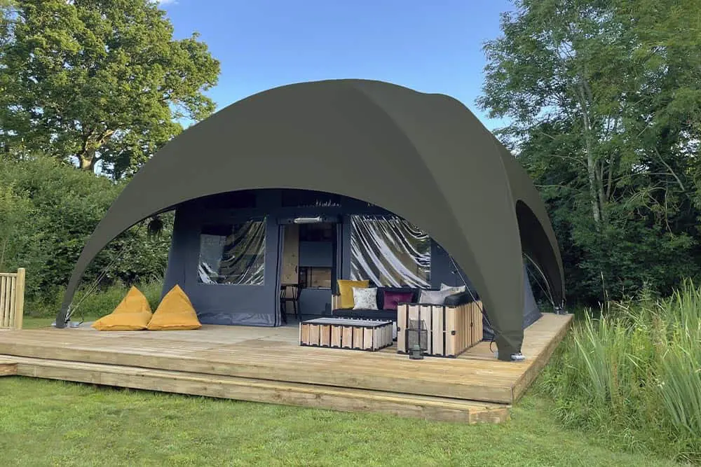 Modern glamping tent with outdoor deck and seating, surrounded by lush green trees, perfect for outdoor adventure.