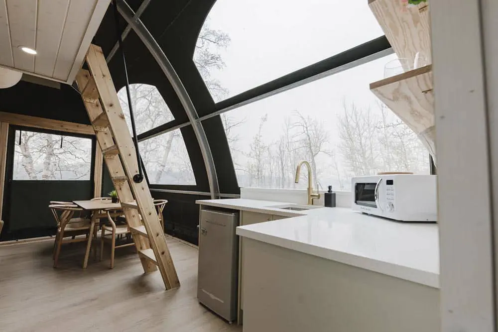 Cozy modern tiny home interior featuring a curved window design, minimalist kitchen with white countertop and wooden furniture, bright and airy space.