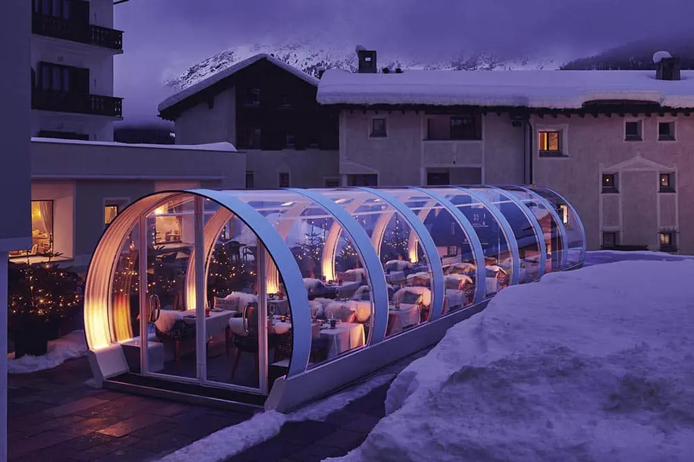 Glass-enclosed winter dining patio with warm lighting and snow-covered surroundings in a mountainous area.