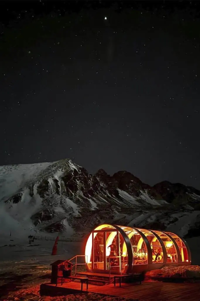 Modern glass igloo accommodation under a starry night sky with snow-covered mountains in the background.