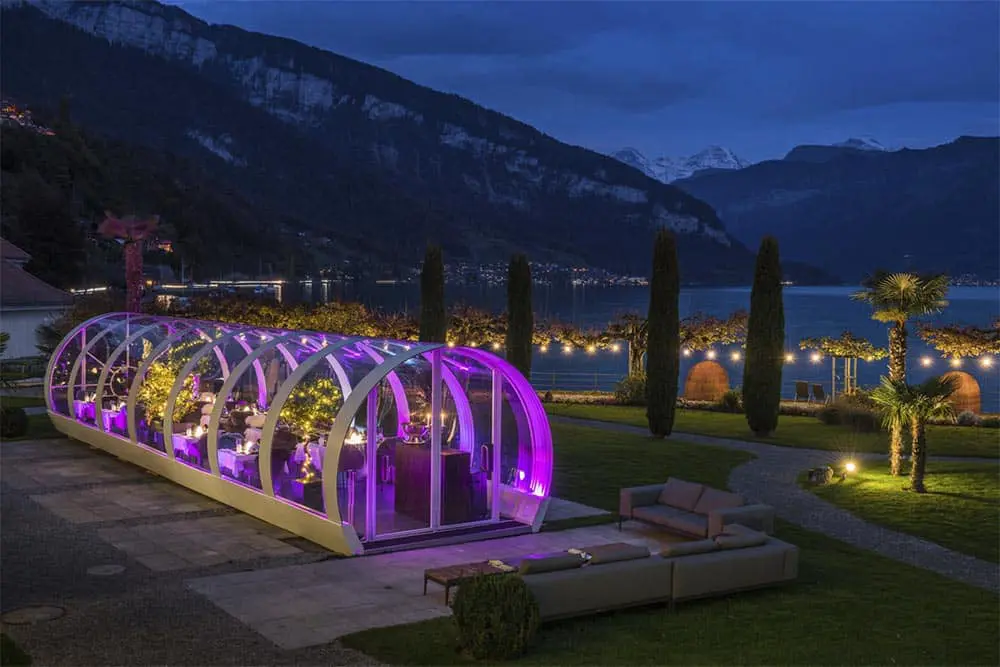 Elegant outdoor dining space with illuminated greenhouse, overlooking a serene lake and mountains, perfect for events and gatherings.