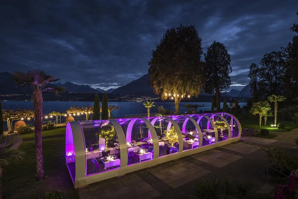 Elegant outdoor dining pod at night with scenic lake and mountain view, illuminated by vibrant purple lighting.