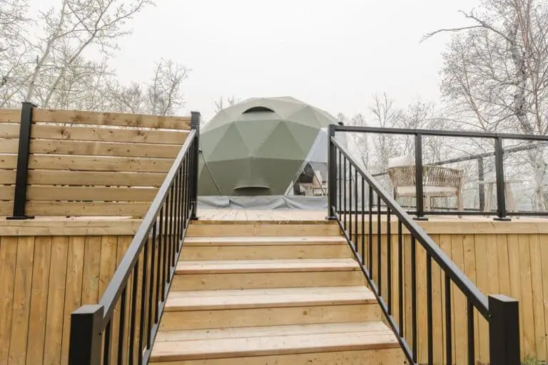 Large wooden deck with black metal railing, featuring a modern geodesic dome outdoor tent, perfect for glamping or outdoor relaxation, set against a backdrop of leafless trees in a natural setting.