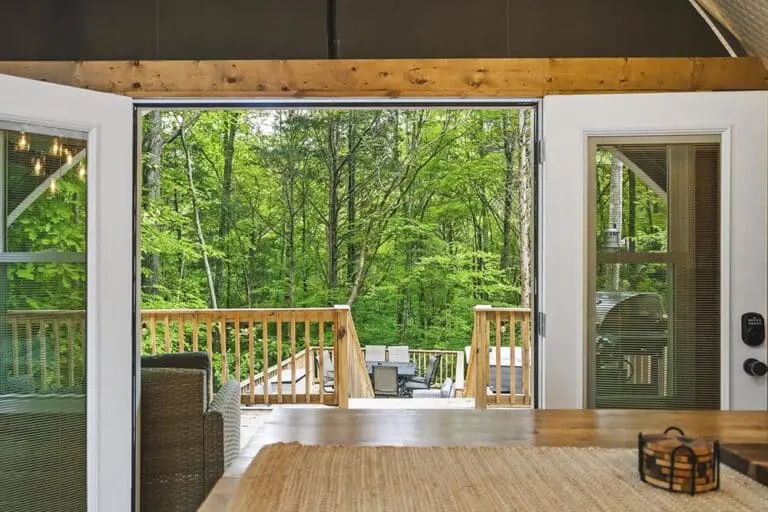 Vast green forest view from modern living room with open door to deck, outdoor seating, natural light, and eco-friendly design.