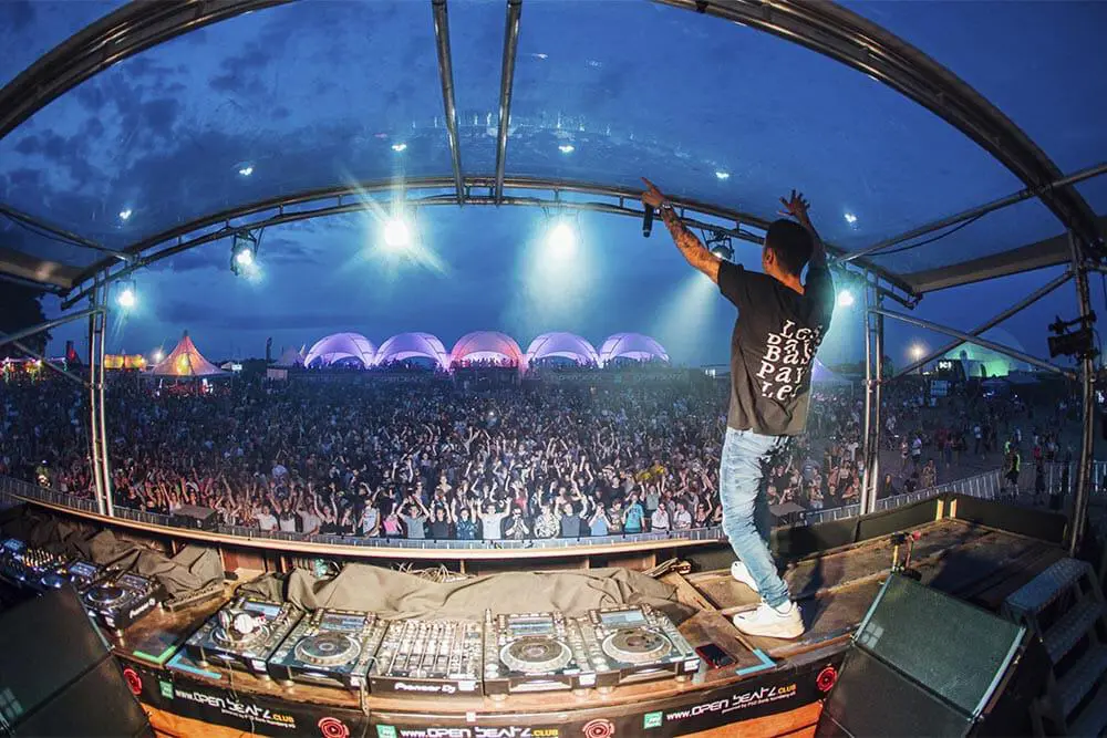 Vast electronic music festival with DJ performing on stage and large crowd in front, illuminated by colorful lights.