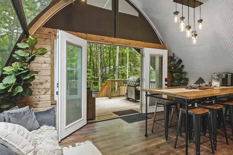 Cozy indoor space opening to a lush outdoor deck with greenery and a barbecue grill, with modern lighting and wooden accents.