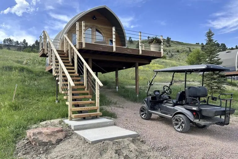Modern tiny house on elevated wooden deck with outdoor stairs and golf cart parked nearby, surrounded by lush green landscape.