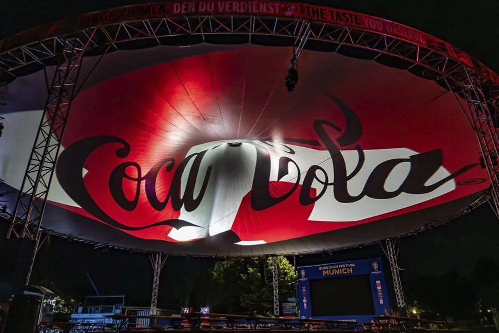 Coca-Cola logo projection on large outdoor stage at nighttime event.