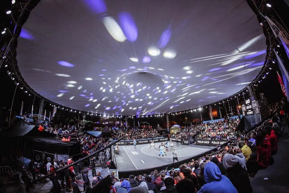 Vibrant indoor 3x3 basketball game under a large illuminated dome with cheering audience.