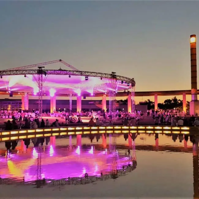 Vibrant outdoor event with stage lighting and audience at dusk, featuring scenic water reflection.