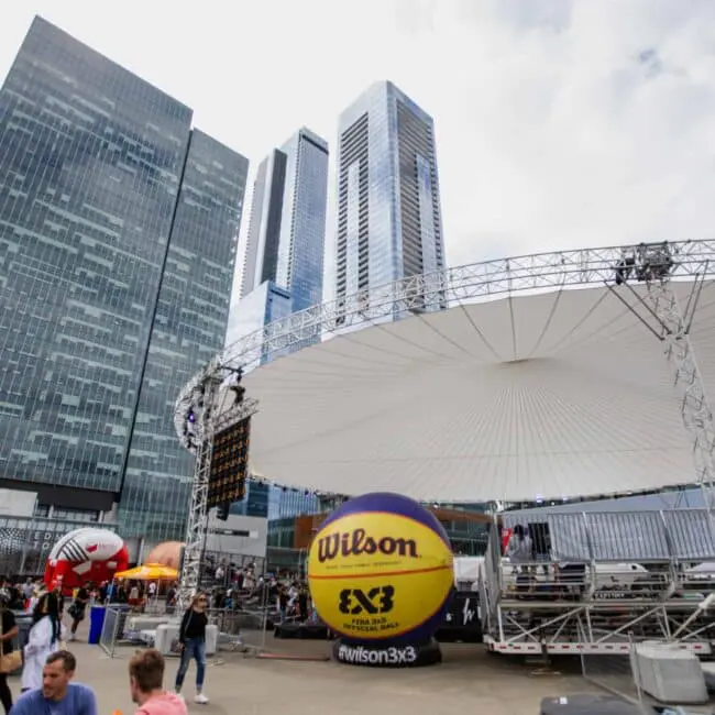 Large outdoor event setup with Wilson 3x3 basketball, high-rise office buildings in background, city outdoor gathering, modern urban venue.