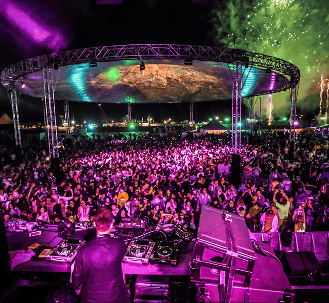 Vibrant outdoor concert with DJ, colorful lighting, fireworks, and lively crowd at night.