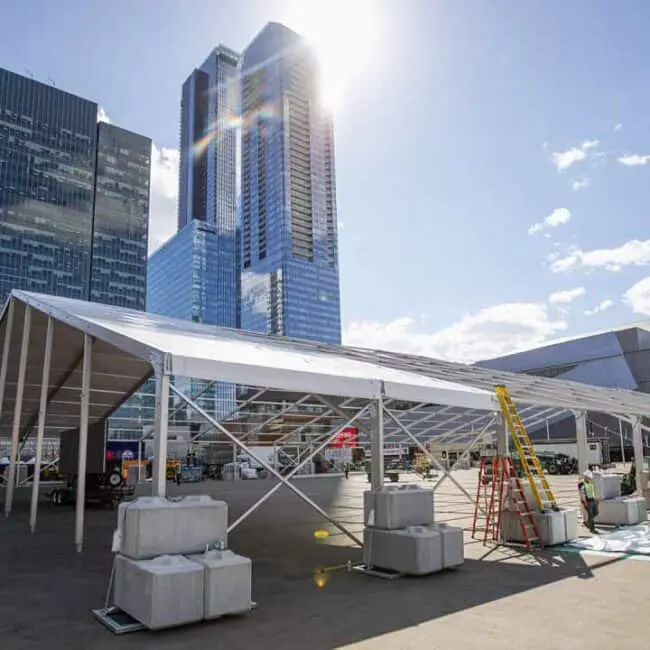 Temporary outdoor event tent being assembled with workers in an urban cityscape, high-rise buildings in the background.