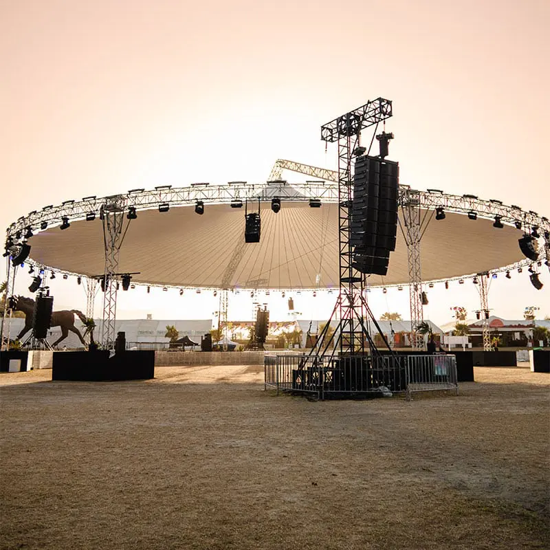 Large outdoor event stage with lighting and sound equipment, under a wide canopy at sunset.