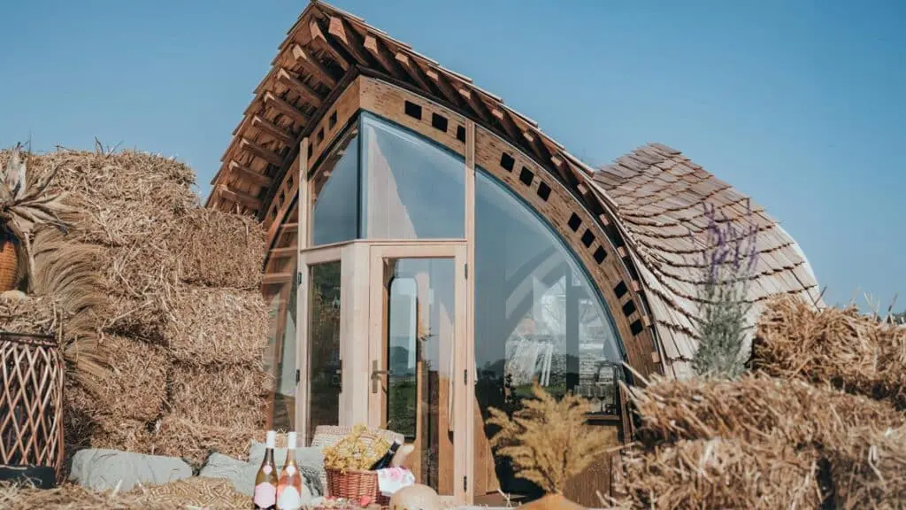 Vegan eco-friendly tiny house with curved wooden roof and glass front door, surrounded by hay bales and rustic decorations.