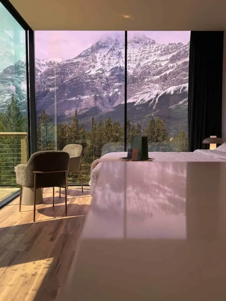Snow-capped mountain view from modern hotel room window, cozy interior with a bed, chairs, and scenic landscape, highlighting luxury accommodations and picturesque nature scenes.