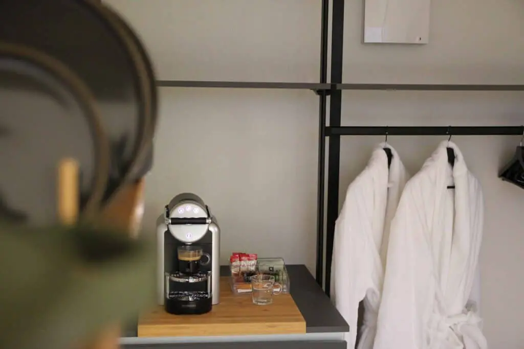 Freshly brewed coffee in a modern hotel room with robes hanging, coffee station with Nespresso machine, sugar, and glasses on a wooden surface, showcasing hospitality essentials for guest comfort.