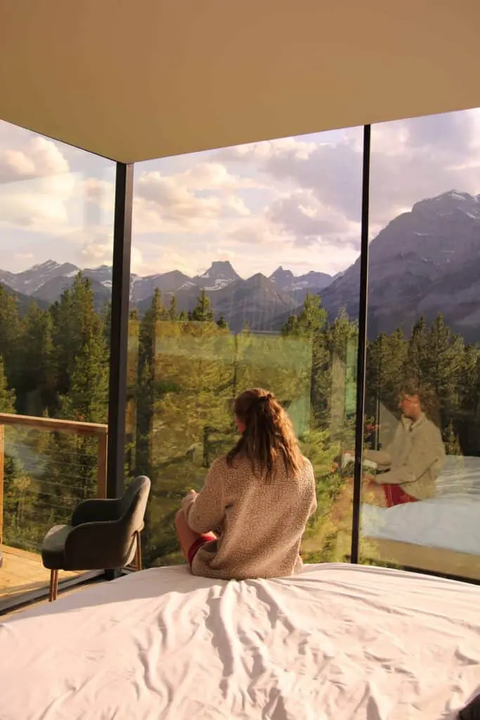 Serene woman sitting on a bed with mountain views through floor-to-ceiling glass windows, highlighting luxury hospitality, nature, and scenic landscapes.