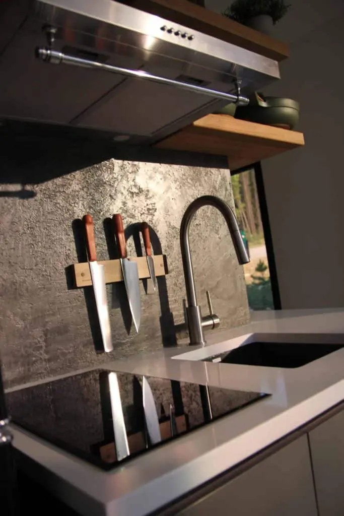 Knife set with wooden handles on magnetic strip in modern kitchen, stainless steel faucet, gray textured countertop, natural light from large window, sleek kitchen design, home cooking essentials, kitchen organization, stylish and functional kitchen decor, high-quality kitchen tools, culinary equipment for cooking and meal prep.