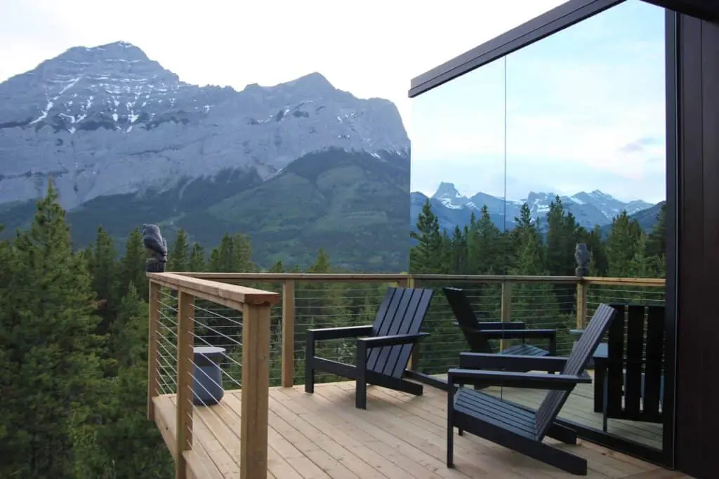 A scenic mountain view from a modern wooden balcony with black chairs, overlooking lush green trees and snow-capped peaks, emphasizing luxury outdoor living and breathtaking natural surroundings.