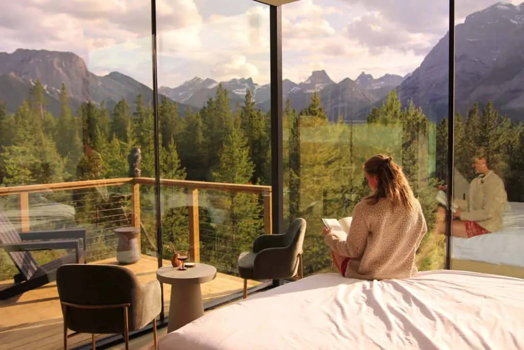 Serene mountain view from a modern hotel room with floor-to-ceiling glass windows, featuring a woman relaxing and reading a book while enjoying the scenic nature landscape and lush forest surroundings.