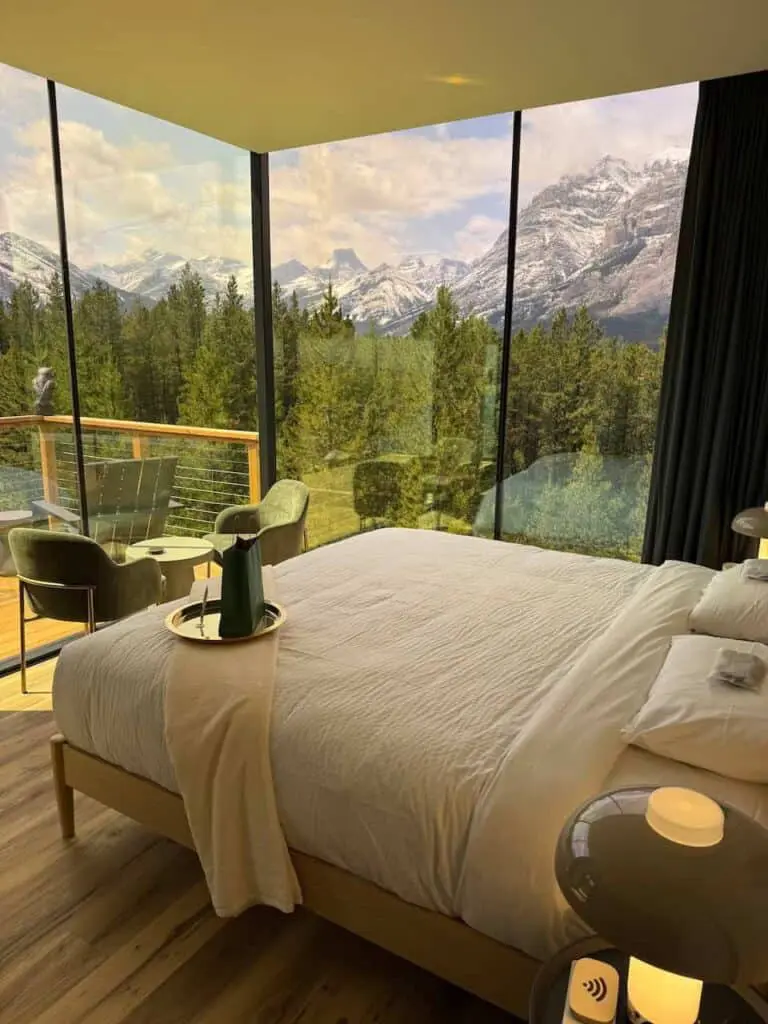 Luxurious mountain view hotel room featuring floor-to-ceiling windows overlooking snow-capped peaks, lush greenery, and modern interior decor with a comfortable bed, balcony seating, and scenic landscape.