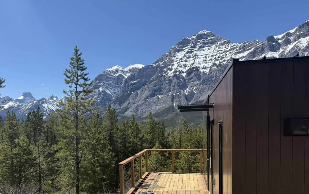 Vivid mountain landscape with snow-capped peaks, lush green pine forest, and modern dark exterior building with wooden deck overlooking scenic views.