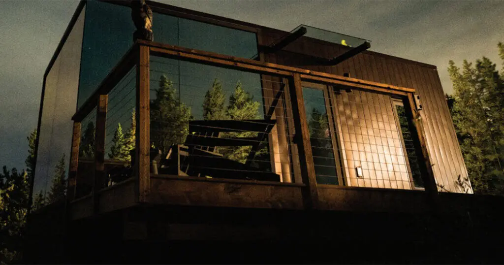 Contemporary glass house featuring large windows, wooden exterior, and surrounding lush greenery. Nighttime setting emphasizes modern design and natural integration.
