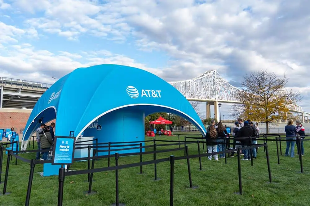 A large blue AT&T branded tent set up outdoors with people queuing, informational signage, and a nearby bridge. Perfect for showcasing technology, community outreach, and customer engagement events.