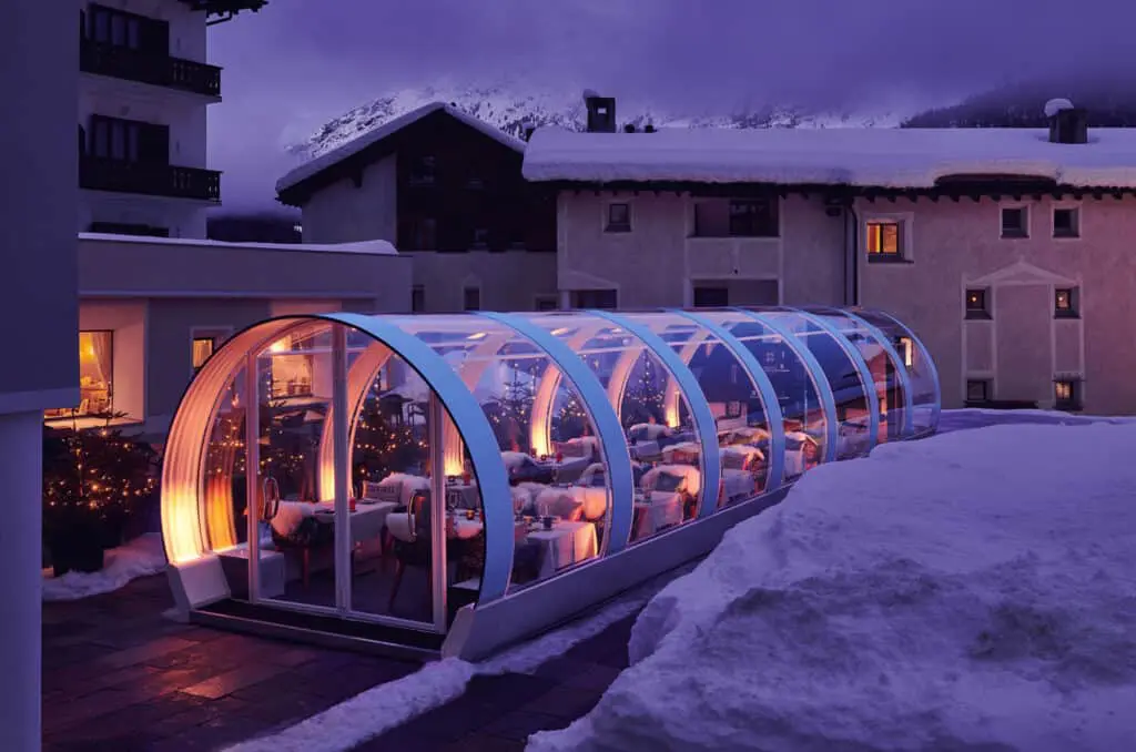 Modern glass winter garden conservatory with warm interior lighting, situated outdoors in a snowy mountain setting, perfect for relaxing and enjoying winter views.