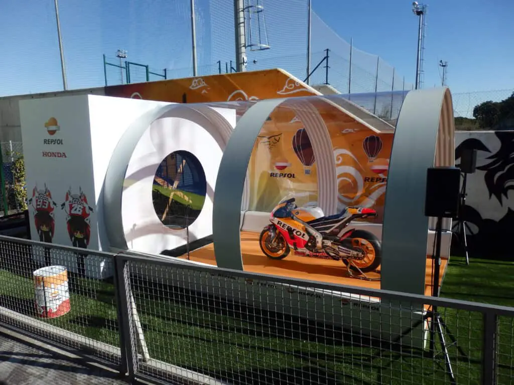 Vivid Repsol Honda motorcycle showcased in a modern exhibition setup with dynamic circular structures, outdoor sports venue background, and branding elements.