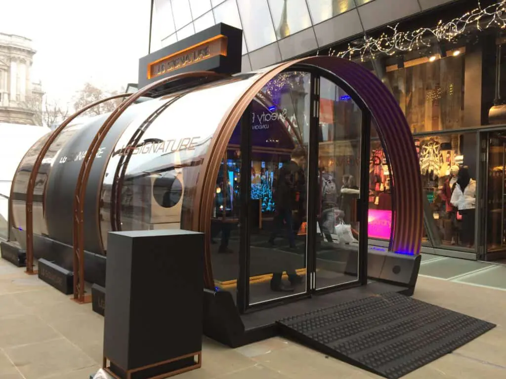 A futuristic, transparent pop-up activation booth with curved design, located outside a retail store, attracting passersby for brand engagement.