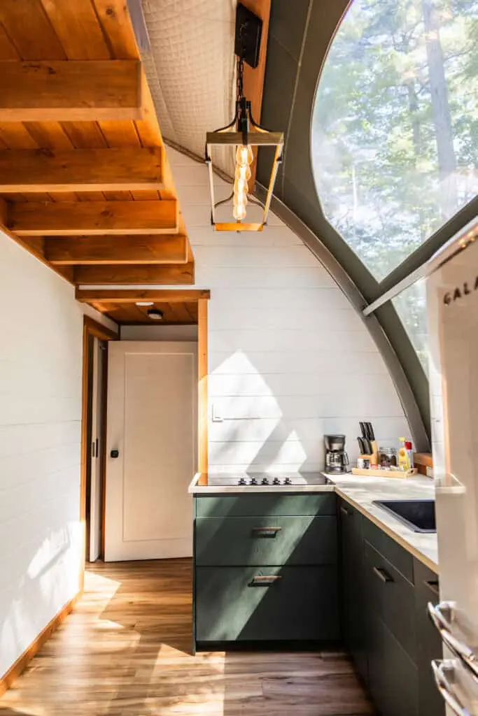 Bright, airy kitchen with wooden ceiling beams, green cabinets, and a large curved window overlooking trees.
