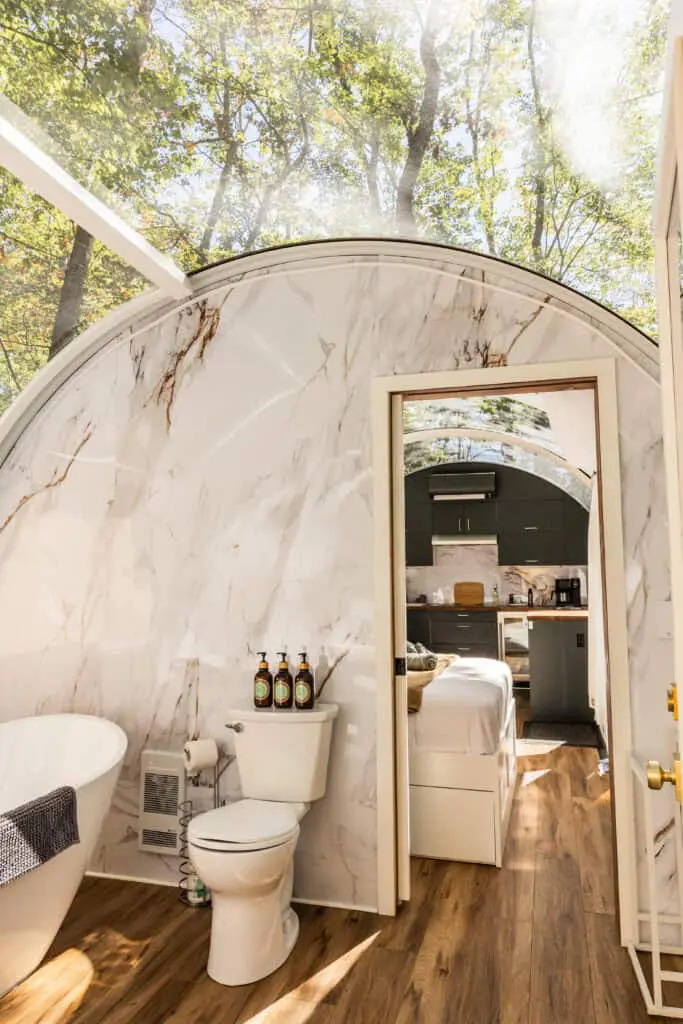 Interior view of a cozy tiny house with marble walls, a bathtub, and a small kitchen area, surrounded by trees and natural sunlight.