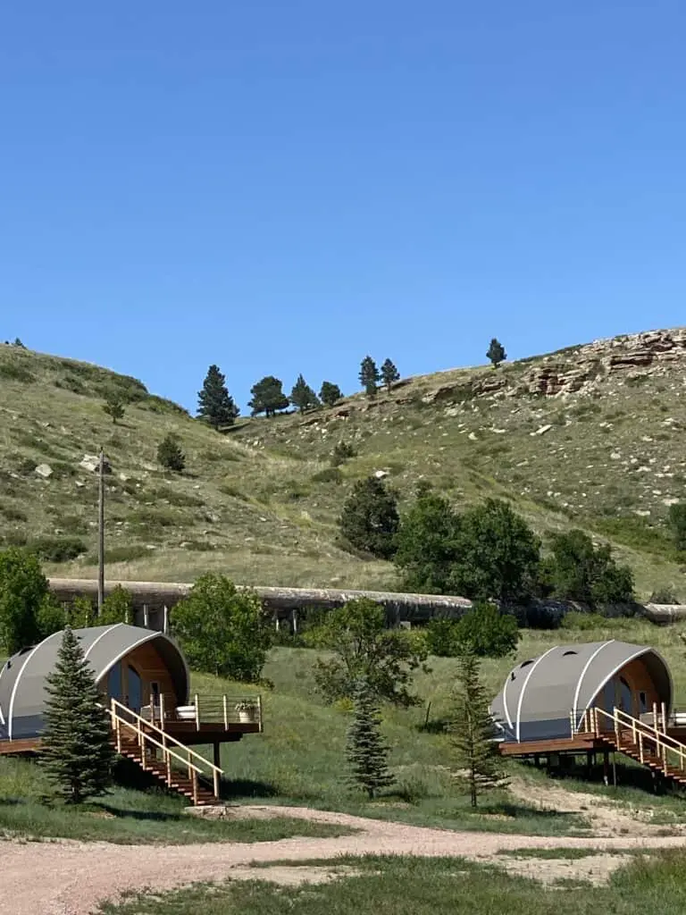 Secluded eco-friendly cabins nestled in a lush green landscape with rolling hills and tall pine trees under a clear blue sky.
