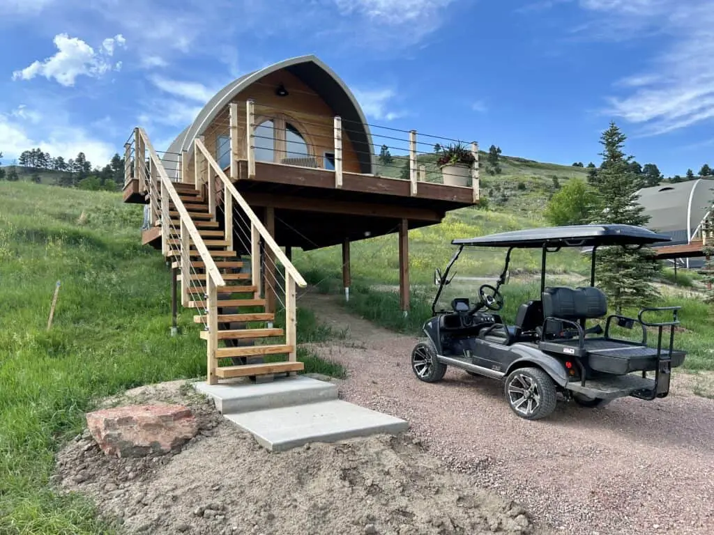 Unique tiny house with curved roof, elevated on stilts, featuring a spacious deck and outdoor stairs.