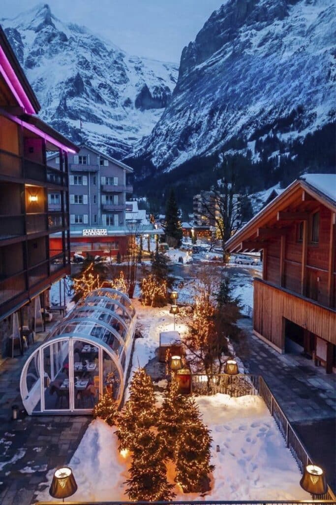 Snow-covered mountain village with illuminated Christmas trees and cozy wooden buildings.