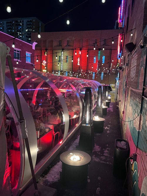 Covered outdoor dining area with colorful string lights and heaters at night, adjacent to a decorated brick wall.