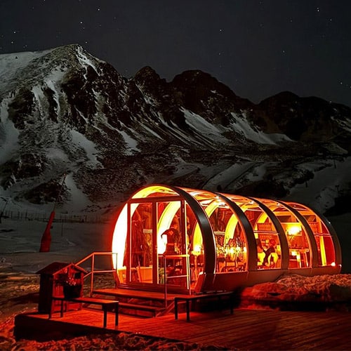Modular glass ice bar in snowy mountain landscape at night, illuminated with warm lighting.