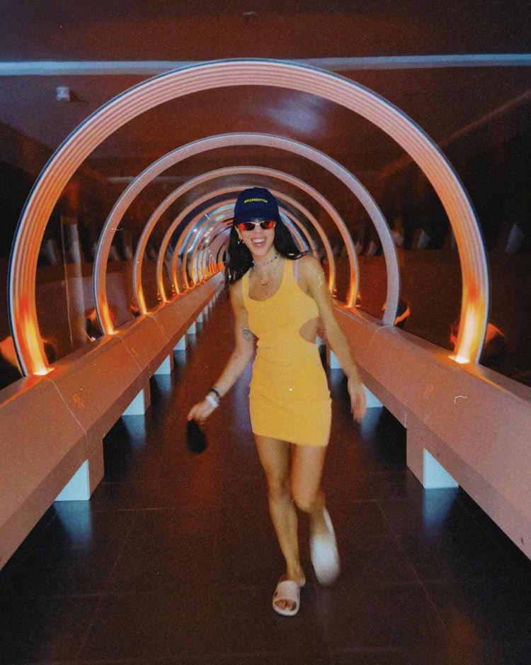Sunny woman in yellow dress exploring futuristic tunnel illuminated with neon lights.