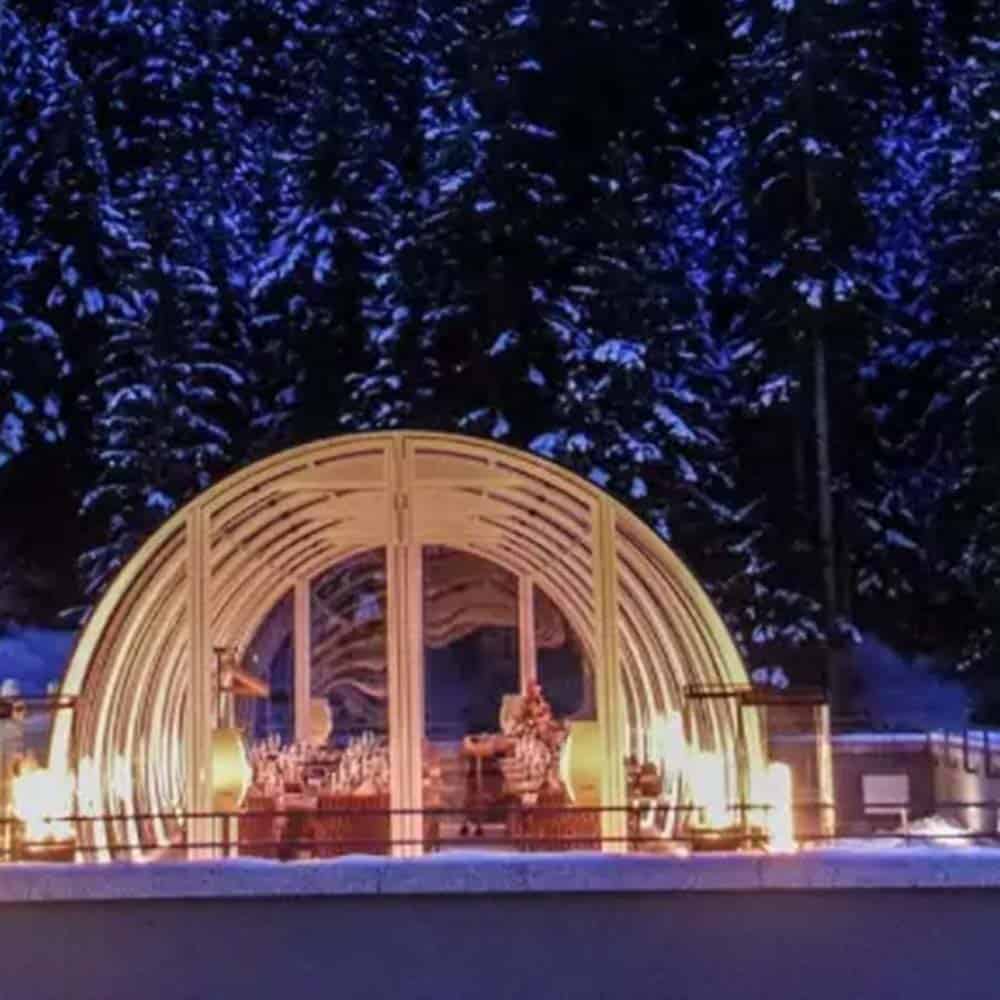 Warm wooden arch structure illuminated at night against snowy evergreen forest, winter outdoor display, festive holiday ambiance.
