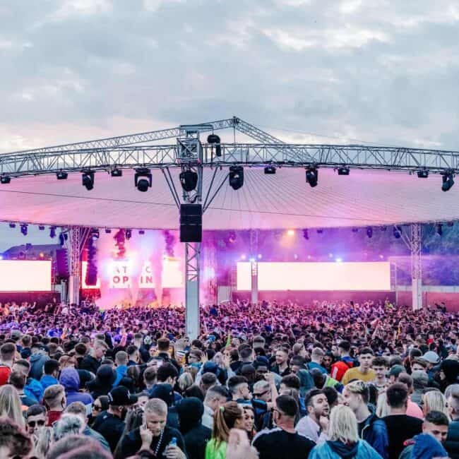 Vibrant outdoor music festival with a large crowd, stage with lights and screens, lively atmosphere, and night sky backdrop.