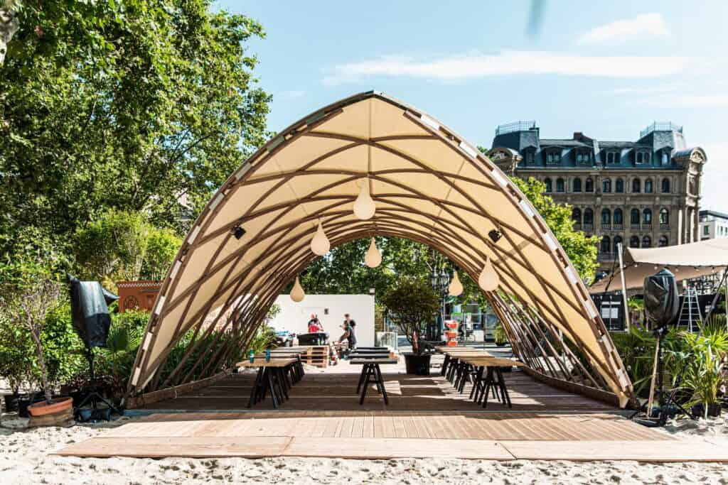 Modern outdoor event space with a stylish wooden arched canopy, surrounded by greenery and urban buildings, for events and gatherings.