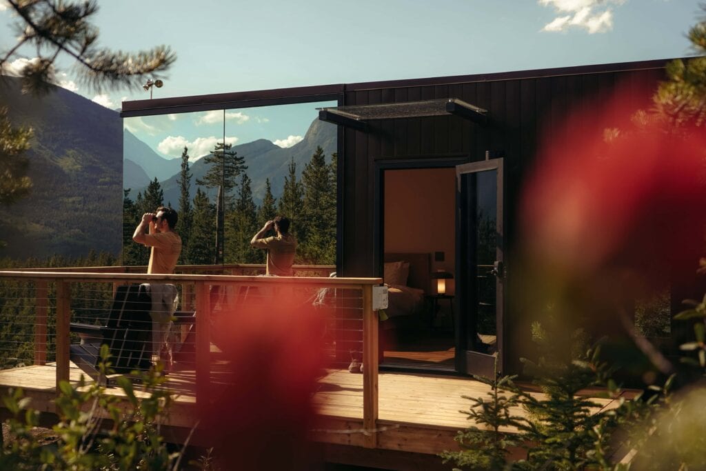 Modern mountain cabin with large glass windows and a spacious wooden deck, surrounded by lush trees and scenic mountain views, perfect for outdoor relaxation and nature escapes.