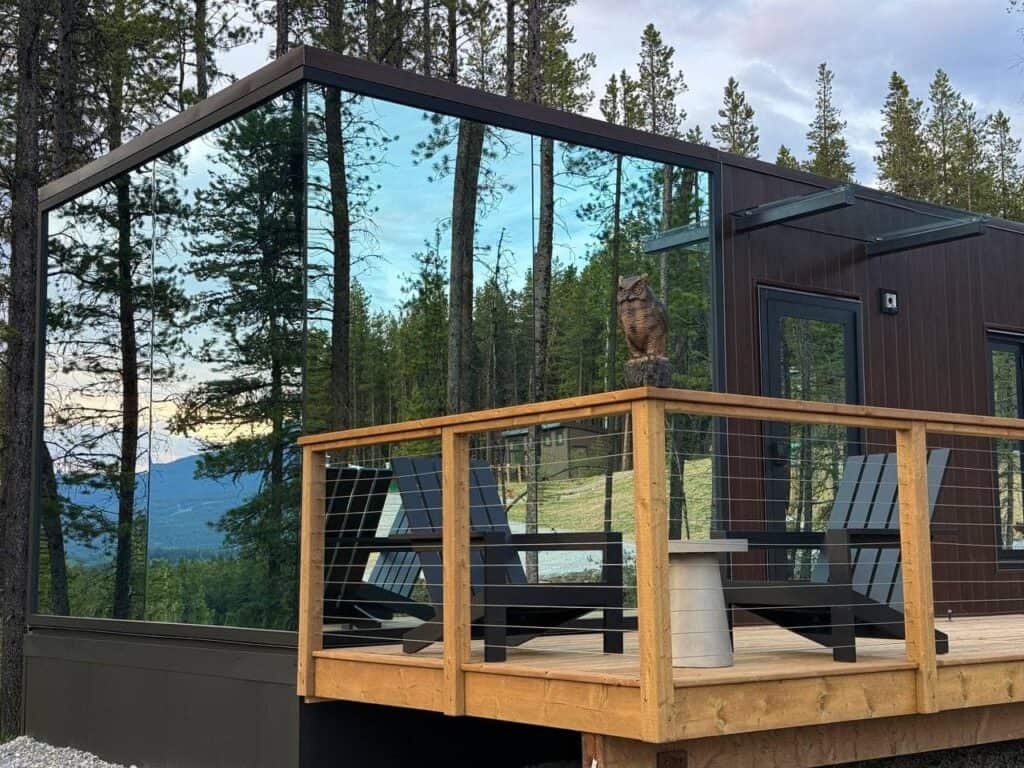 Modern tiny home with large glass windows overlooking a forested mountain landscape, featuring an outdoor wooden deck with Adirondack chairs and an owl sculpture for nature-inspired living.