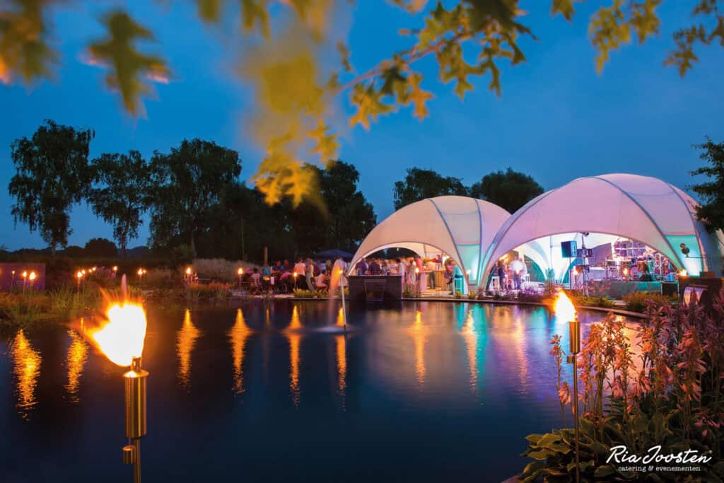A lively outdoor event featuring illuminated tents and fire torches by a water body during dusk, showcasing entertainment and social gathering in an industrial-inspired setting.