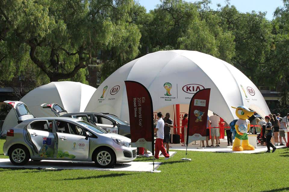 Interactive brand activation event with a large white dome tent, cars, and mascot at an outdoor festival. Engaging activities for visitors under sunny skies.
