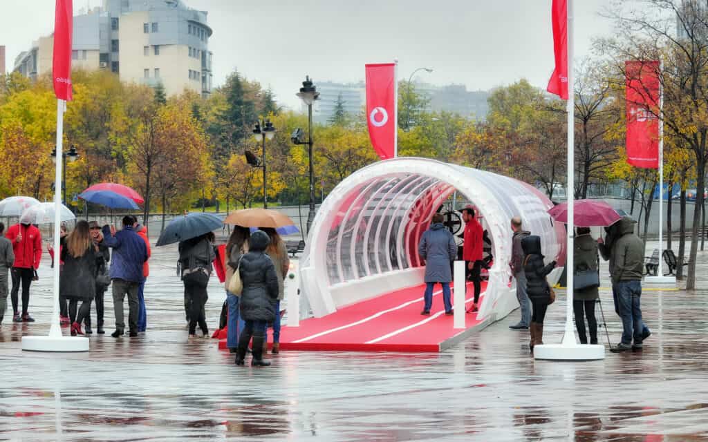 People gather around a modern, transparent event tunnel with red accents during an outdoor experiential activation on a rainy day.